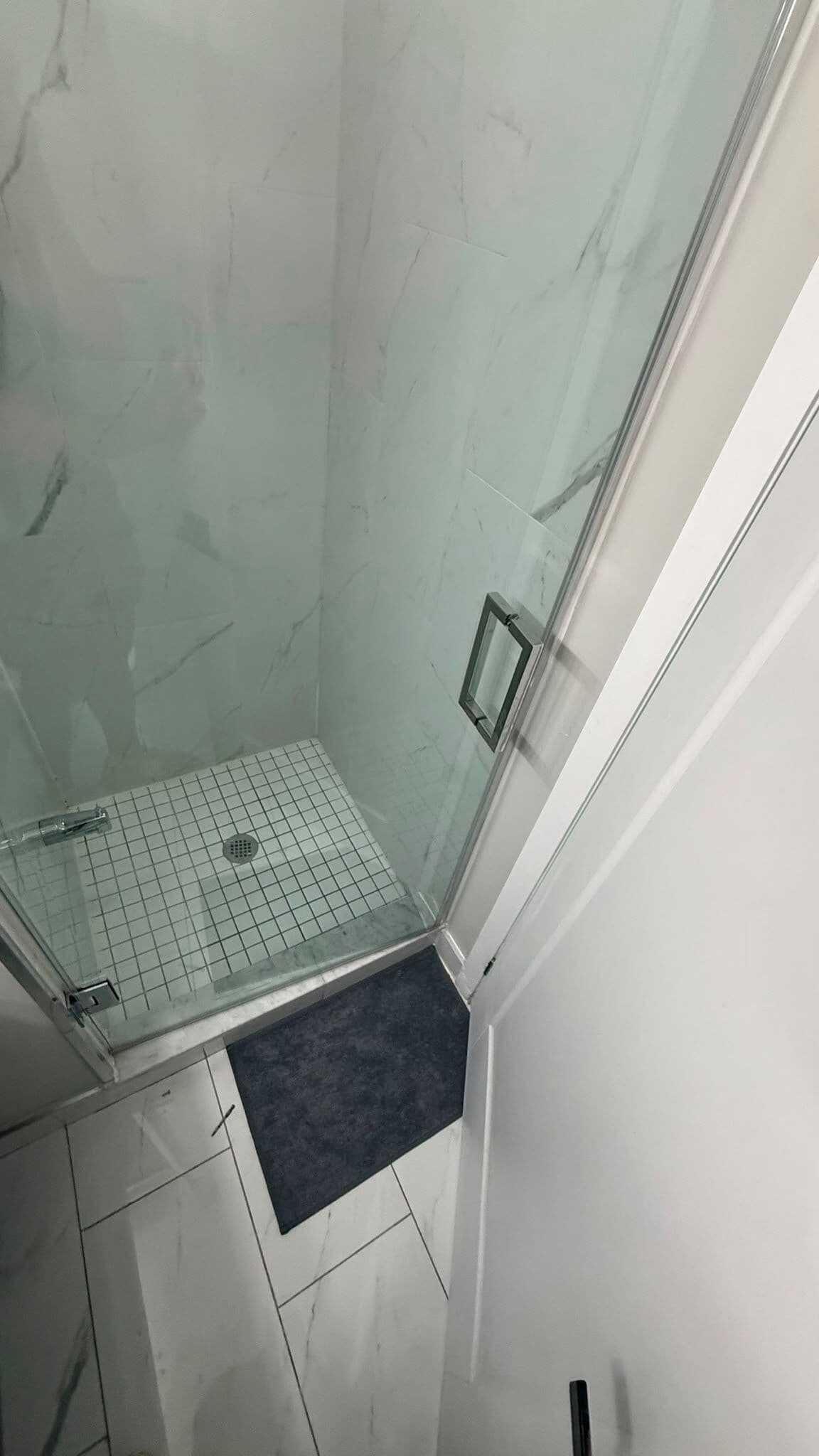 A shower with a glass door, a tiled floor, and a small mat.