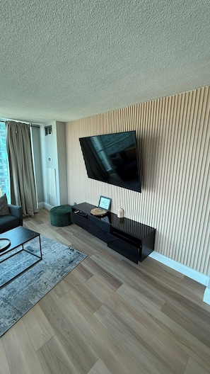 A modern living room with a flat-screen TV mounted on a wooden panel wall, a dark sofa, a coffee table, and a green ottoman.