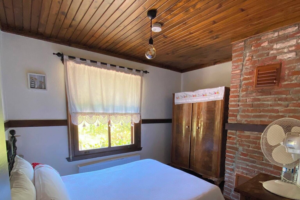 A bedroom with a wooden ceiling, a bed with white bedding, a window with a curtain, and a brick wall.