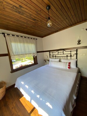 A bedroom with a wooden ceiling, a bed with white bedding, a window with a view of greenery, and a bedside table with a lamp. - Papuli Köy Evi (Karamürsel)