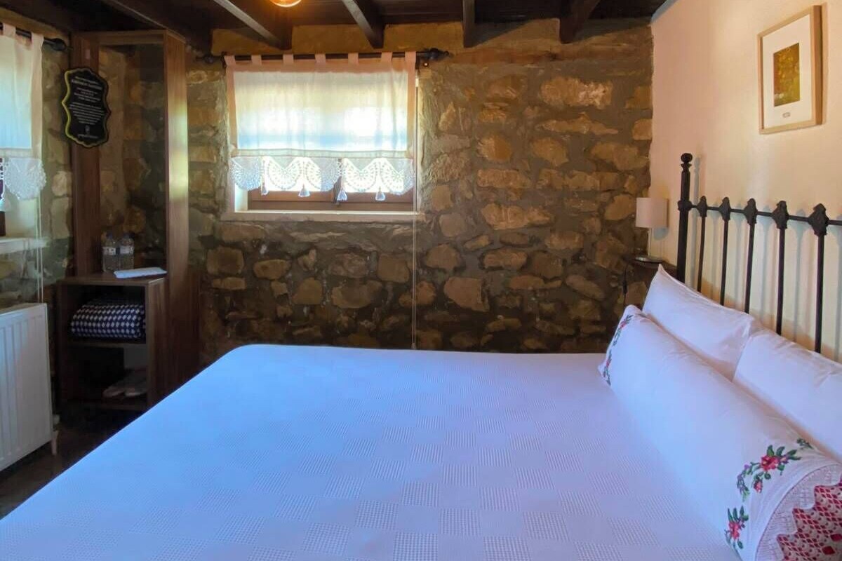 A bedroom with a stone wall, a bed with white bedding, a window with lace curtains, and a wooden ceiling.