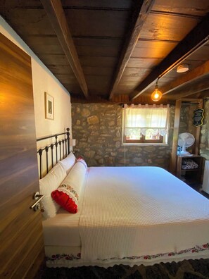 A bedroom with a stone wall, a bed with a white bedspread, a red pillow, a wooden ceiling, and a window with a curtain. - Papuli Köy Evi (Karamürsel)