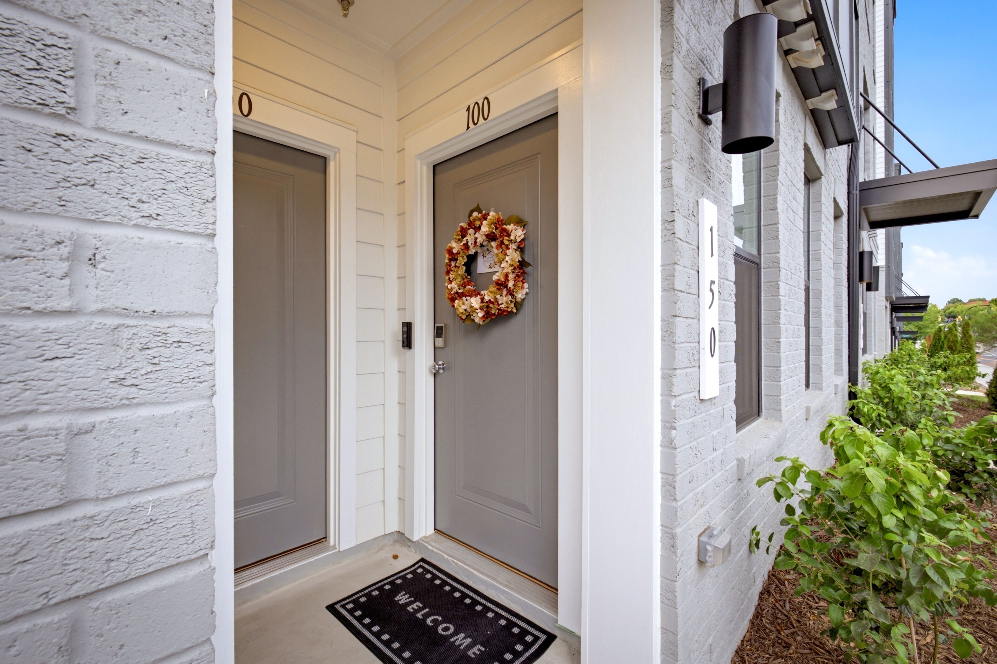 A white building with two doors, each marked with a number, a welcome mat, and a wreath on the door.
