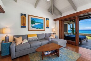 A living room with a grey sofa, a wooden coffee table, and a view of the ocean through a sliding door. - Island Time Retreat by Gather: Oceanfront Access (Kamuela)