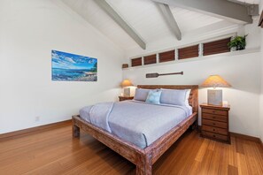 A bedroom with a wooden bed, two bedside tables with lamps, a wall-mounted shelf with plants, and a painting of a beach scene. - Island Time Retreat by Gather: Oceanfront Access (Kamuela)
