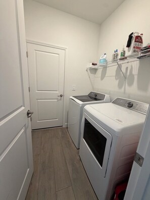 A laundry room with a washing machine and dryer, a door, and a shelf with cleaning supplies. - Gated Babcock Ranch Home - Pool, Spa, Sauna & Lake View (Punta Gorda)