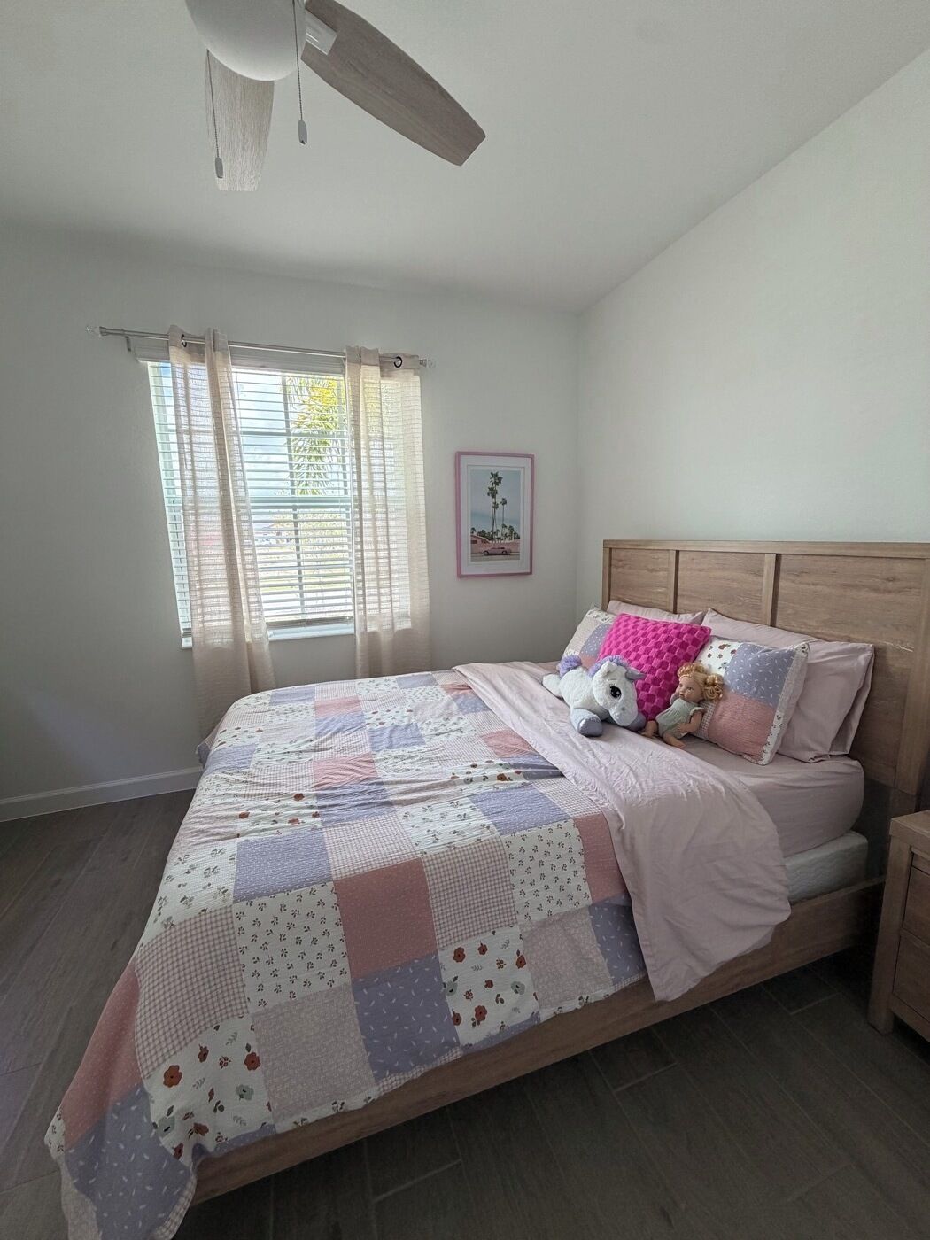 A bedroom with a wooden headboard, a quilted bedspread, a window with sheer curtains, and a framed picture on the wall.