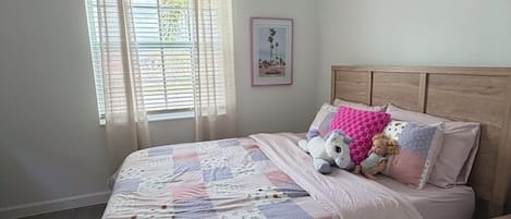 Un dormitorio con cabecera de madera, colcha acolchada, ventana con cortinas de gasa y un cuadro enmarcado en la pared.