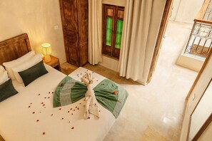 A hotel room with a bed, a nightstand, a lamp, a window with curtains, and a view of greenery outside. - Vrbo Property (Marrakech)