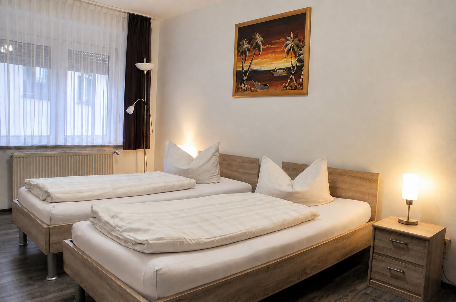 Ein Hotelzimmer mit zwei Betten, einem Nachttisch mit Lampe, einem Bild an der Wand und einem Fenster mit Vorhängen.