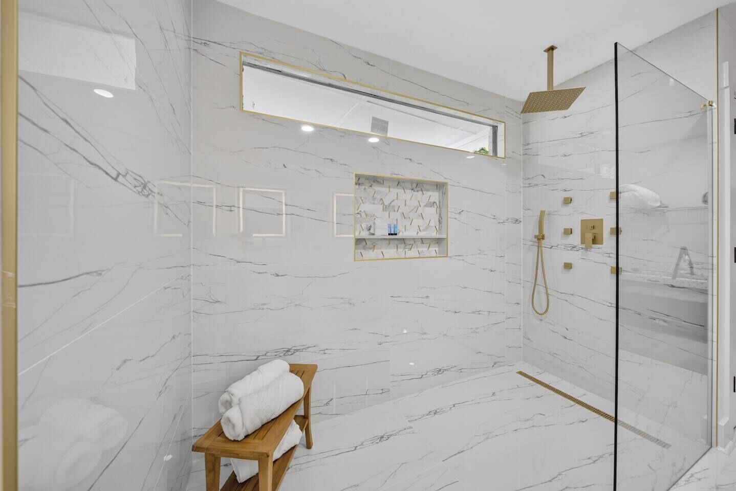 A modern bathroom with marble walls, a glass shower enclosure, and a wooden bench with towels.