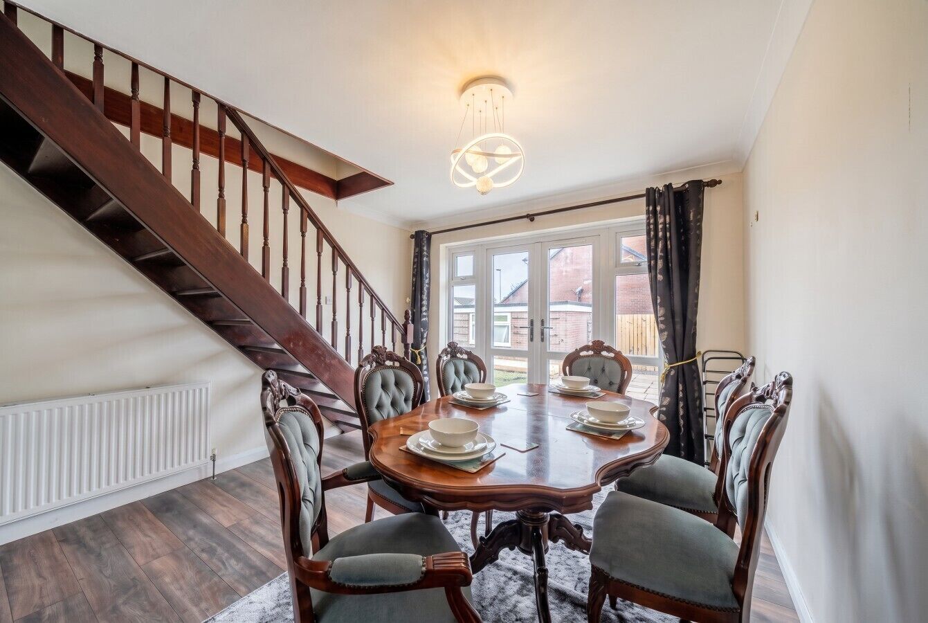 A dining area with a wooden table set for four, surrounded by upholstered chairs, a staircase, and a view through large windows.