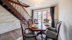 Comedor con una mesa de madera dispuesta para cuatro, sillas tapizadas alrededor, una escalera y vistas amplias a través de grandes ventanales.