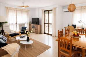 A well-lit living room with a sofa, chairs, a coffee table, and a dining table with chairs. There is a TV, a plant, and a window with curtains.