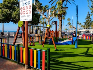A playground with swings and slides, a sign with various prohibitions, and a palm tree in the background.