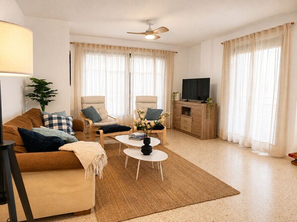 A living room with a sofa, two armchairs, a coffee table, a TV on a wooden cabinet, and a ceiling fan.