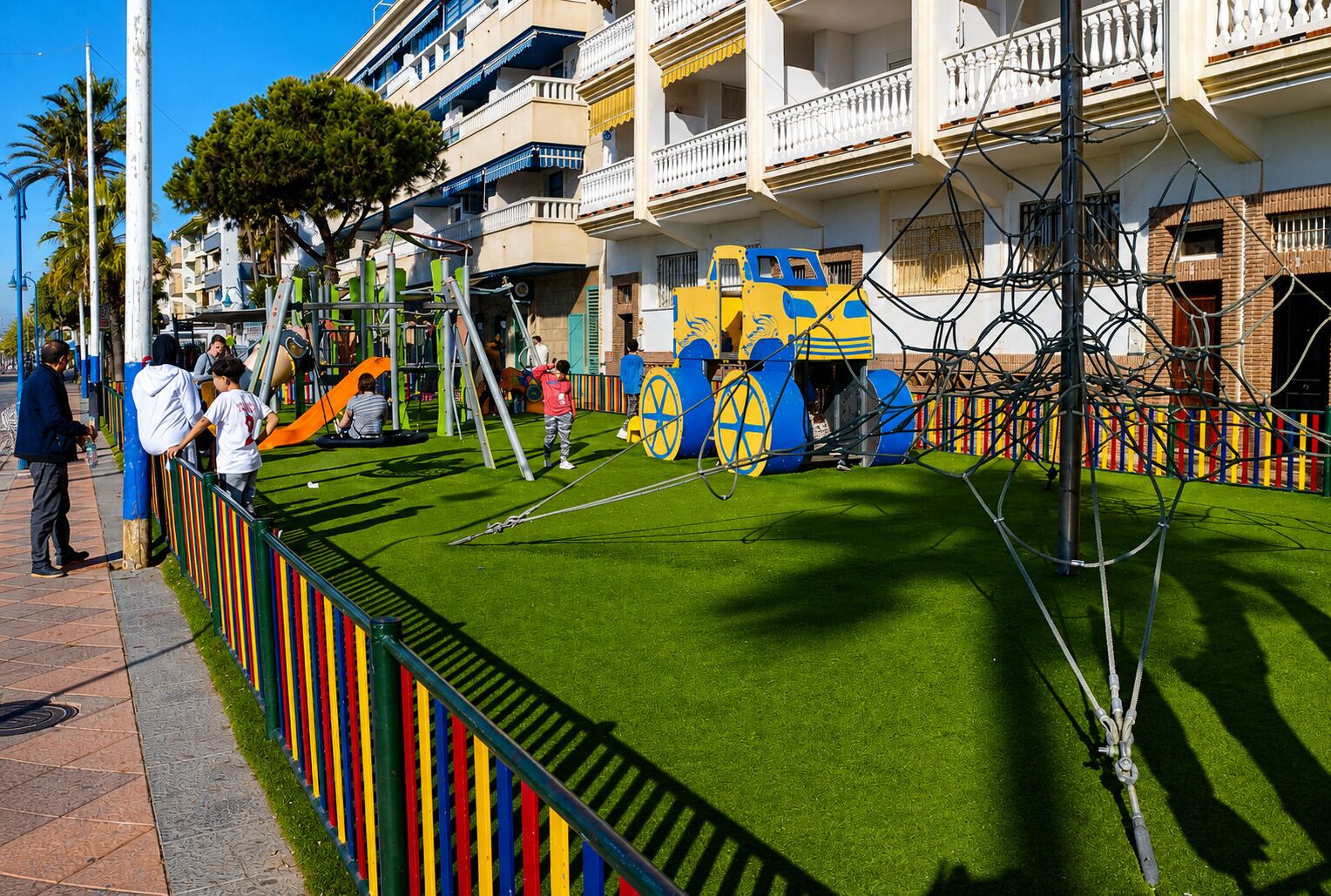 A playground with a green artificial turf surface, colorful play structures, and a fence with vertical stripes.
