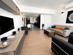 Una sala moderna con un televisor de pantalla plana, una mesa de centro de madera, un sofá con cojines de colores y una zona de comedor al fondo.
