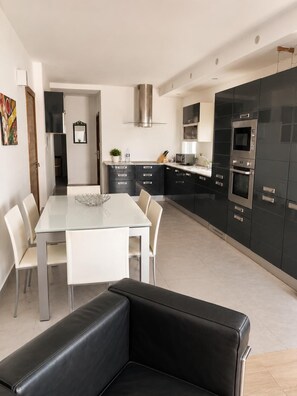 A modern kitchen with black cabinets, stainless steel appliances, and a dining area with a glass table and chairs. - Sliema 2BR Sea View Apartment Qui Si Sana with Terrace & BBQ (Sliema)