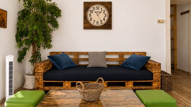 Una habitación con un sofá de madera, dos almohadas, un reloj de pared, una planta en maceta, una cesta de mimbre y un banco verde.