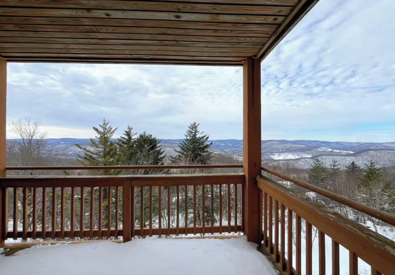 A wooden deck with a snow-covered ground, overlooking a mountainous landscape with trees and a cloudy sky.