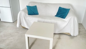Een witte bank met twee blauwe kussens, een kleine witte tafel, en een witte koelkast in een kamer met een effen witte muur en een betegelde vloer.