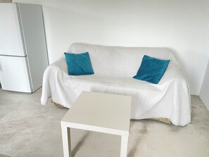A white sofa with two blue cushions, a small white table, and a white refrigerator in a room with a plain white wall and tiled floor. - Préchacq-les-Bains (Préchacq-les-Bains)