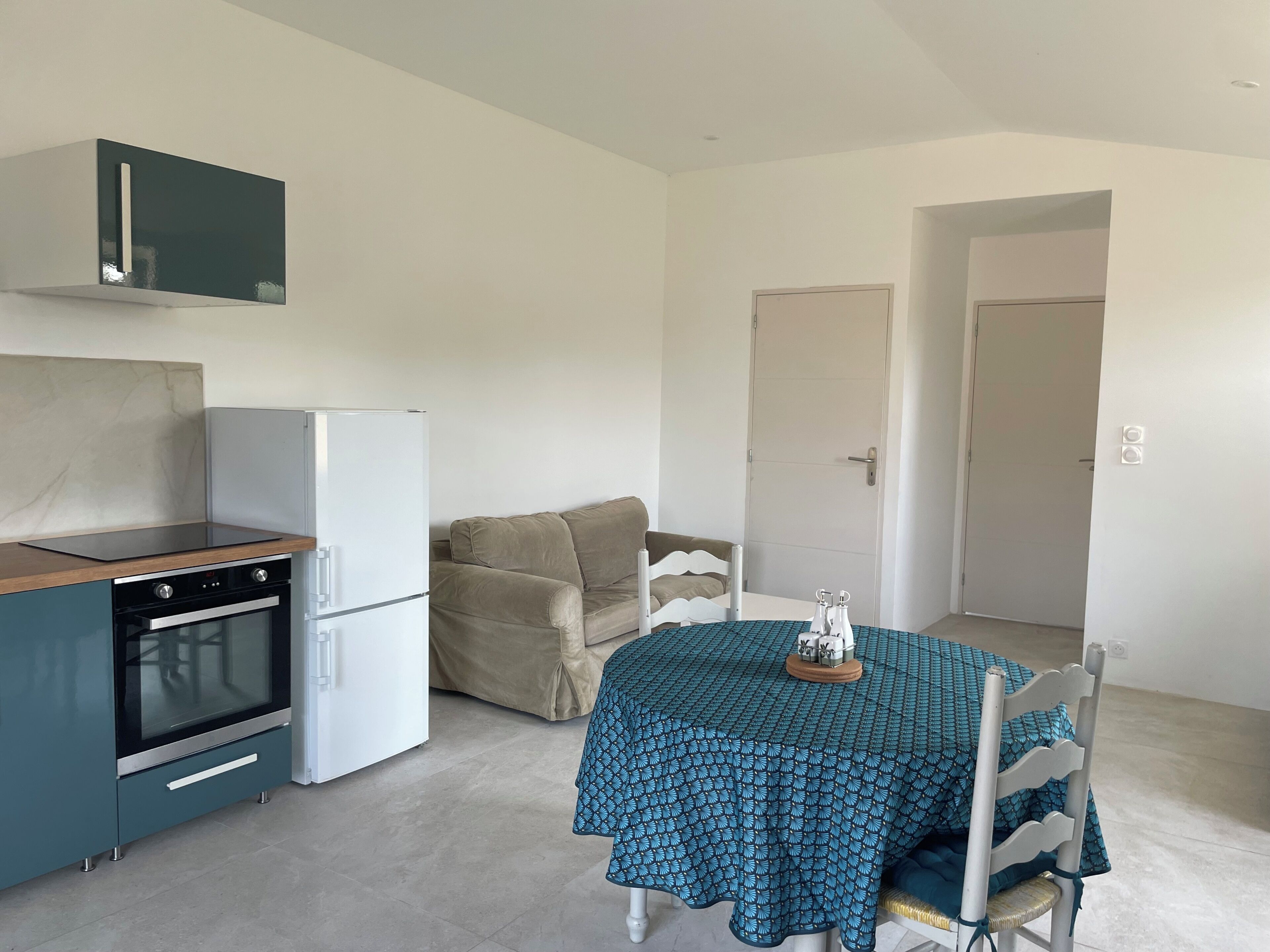 Une cuisine compacte équipée d’une cuisinière, d’un four et d’un réfrigérateur, d’un canapé beige, d’une table à manger recouverte d’une nappe à motifs et d’une porte blanche en arrière-plan.