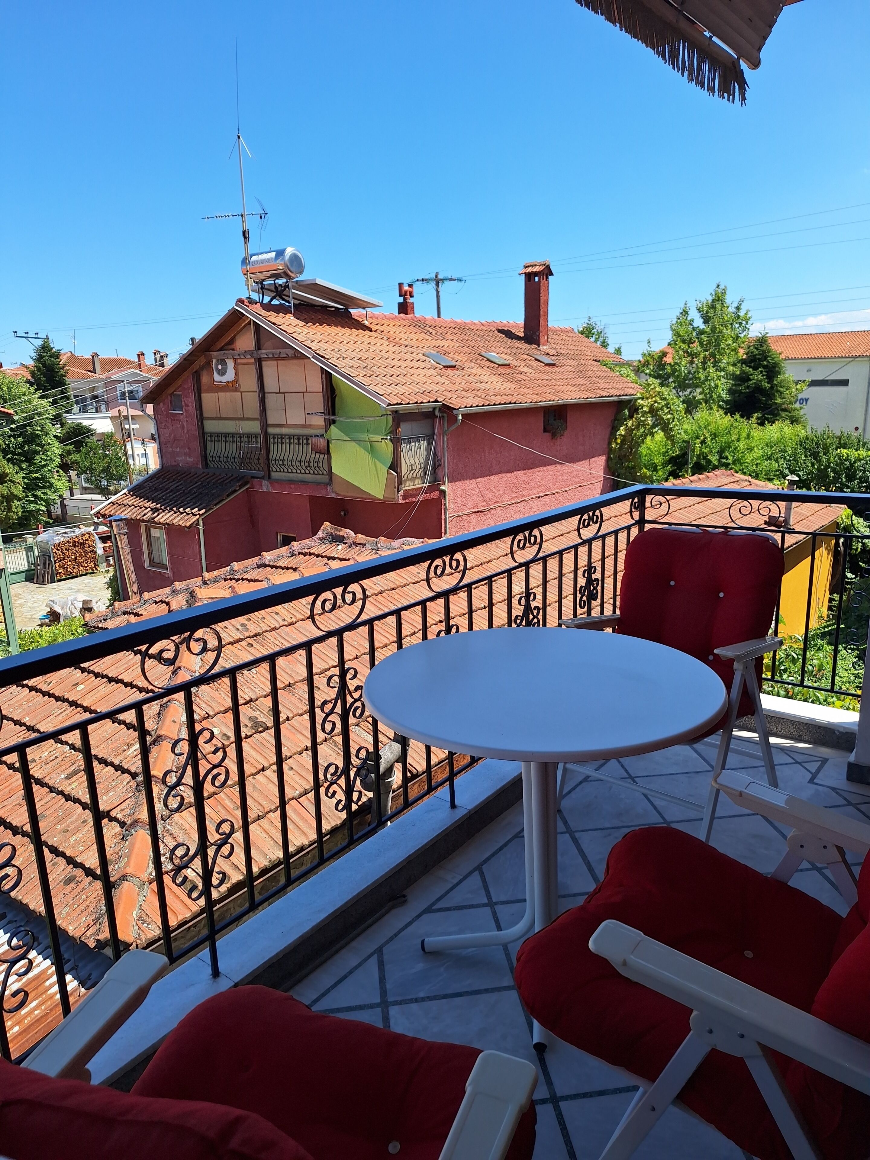 A balcony with a round table and red chairs, overlooking a residential area with terracotta roofs.