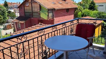 A balcony with a round table and red chairs, overlooking a residential area with terracotta roofs.
