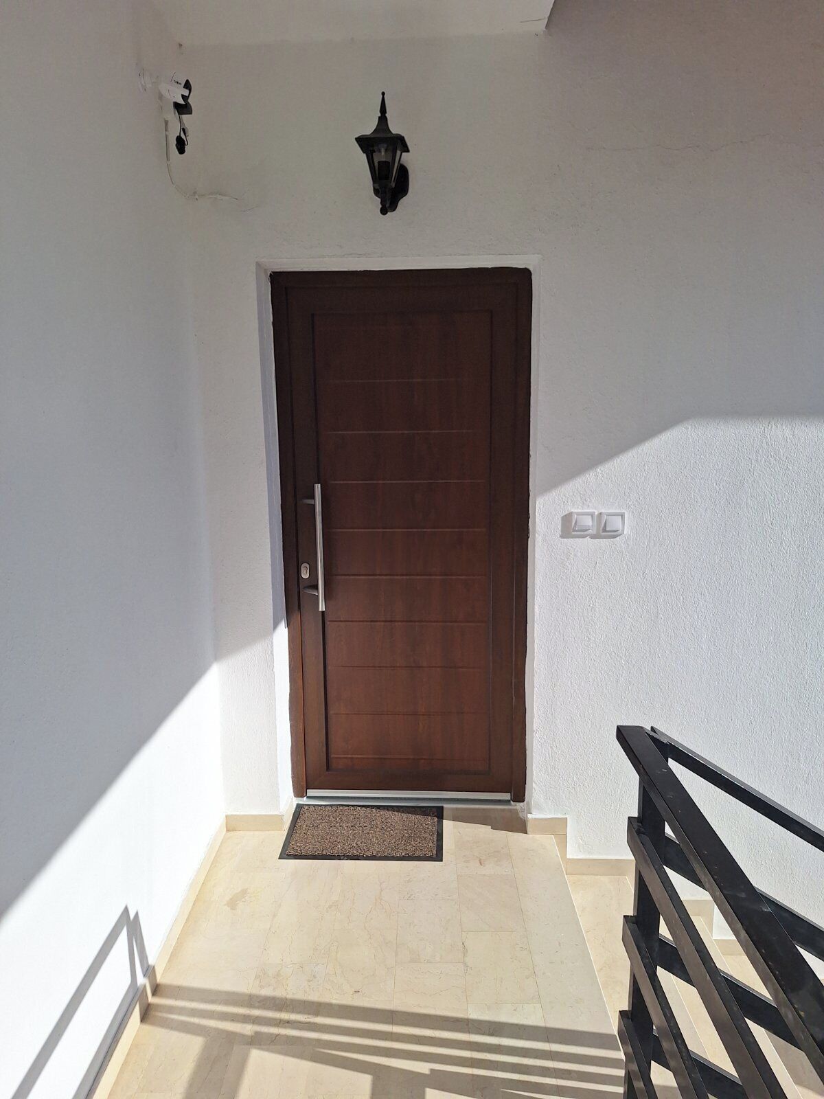A closed wooden door with a metal handle, a small wall-mounted light fixture above it, and a doormat at the entrance.