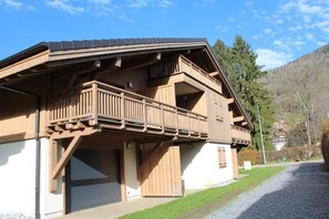 Un bâtiment en bois à deux étages, doté d’un balcon, d’une porte de garage et d’une allée menant à une maison située au fond.