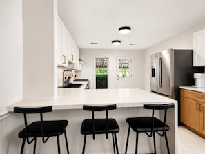 A modern kitchen with a white countertop, three black bar stools, stainless steel appliances, and wooden cabinets. - The Monroe by Willow · Mt Pleasant · 4BR Residence (Washington)