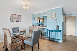 A dining area with a glass table and wicker chairs, a kitchen with wooden cabinets, and a blue accent wall. - The Islander 314 Dolphin Cove (Destin)
