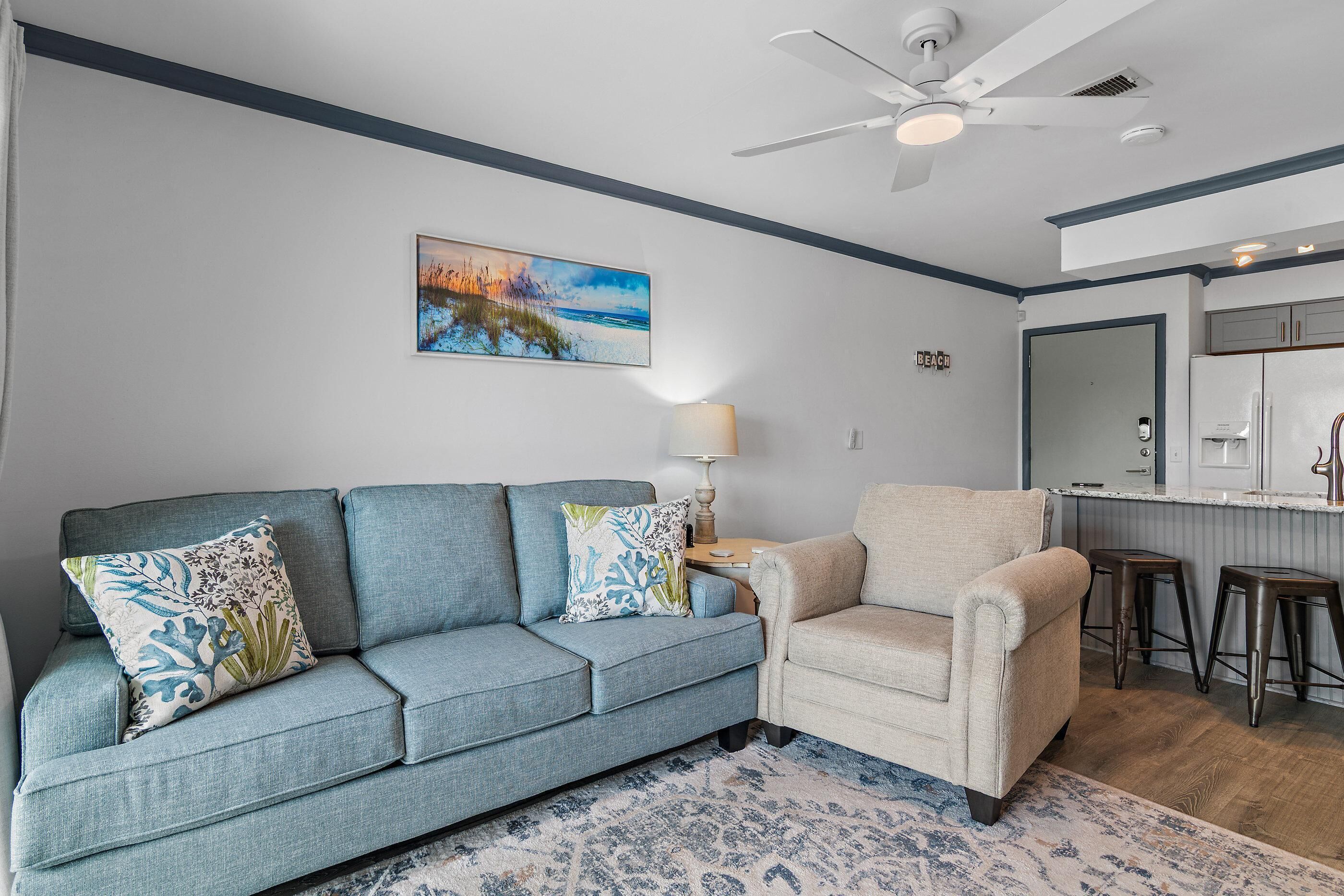 Ein Wohnzimmer mit einem blauen Sofa, einem beigen Sessel, einem Bild einer Strandlandschaft, einem Deckenventilator und einer Küchenzeile mit einem Kühlschrank.