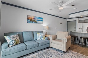 A living room with a blue sofa, a beige armchair, a painting of a beach scene, a ceiling fan, and a kitchen area with a refrigerator. - Lakeview #9 - Paradise By The Gulf (Miramar Beach)