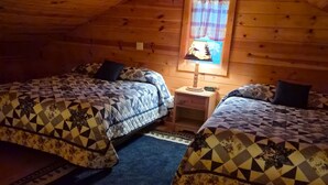 A cozy attic bedroom with two beds, a window with curtains, a ceiling fan, and a nightstand with a lamp.
