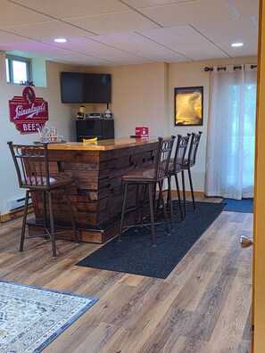 A bar area with a wooden counter, bar stools, a television mounted on the wall, and a framed beer advertisement.