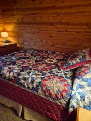 Ein Holzblockhaus-Interieur mit einem Bett, das mit einem Patchwork-Quilt bezogen ist, einem Nachttisch mit Lampe und einem Holz-Kopfteil.
