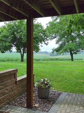 A covered outdoor area with a stone bench, a planter with flowers, and a view of a green field and trees.