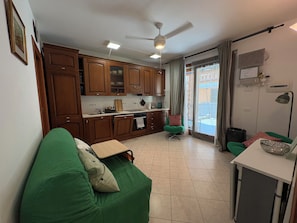 A compact living space with a kitchenette, a green sofa, a dining area, and a view through a sliding door. - Charming apartment with a pool and breathtaking views. (Spinone Al Lago)