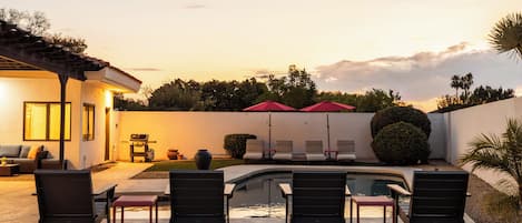 Un área de piscina con sillas de descanso y sombrillas, una parrilla y una casa con patio.