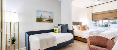 A bedroom with a bed, a desk, a chair, a plant, and a painting on the wall.