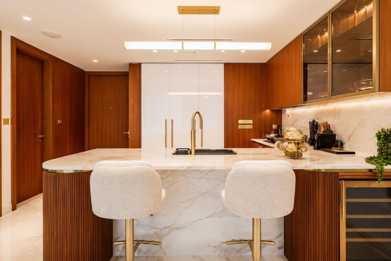 A modern kitchen with a marble countertop, two bar stools, a built-in sink, and wooden cabinetry.