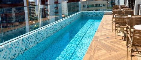 A rooftop swimming pool with a tiled edge, surrounded by wooden decking and wicker furniture.