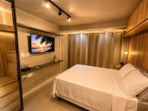 A modern bedroom with a bed, a mounted TV, a shelf with a plant, and a curtain. - Studio na Barra Zn 1403 (Salvador)