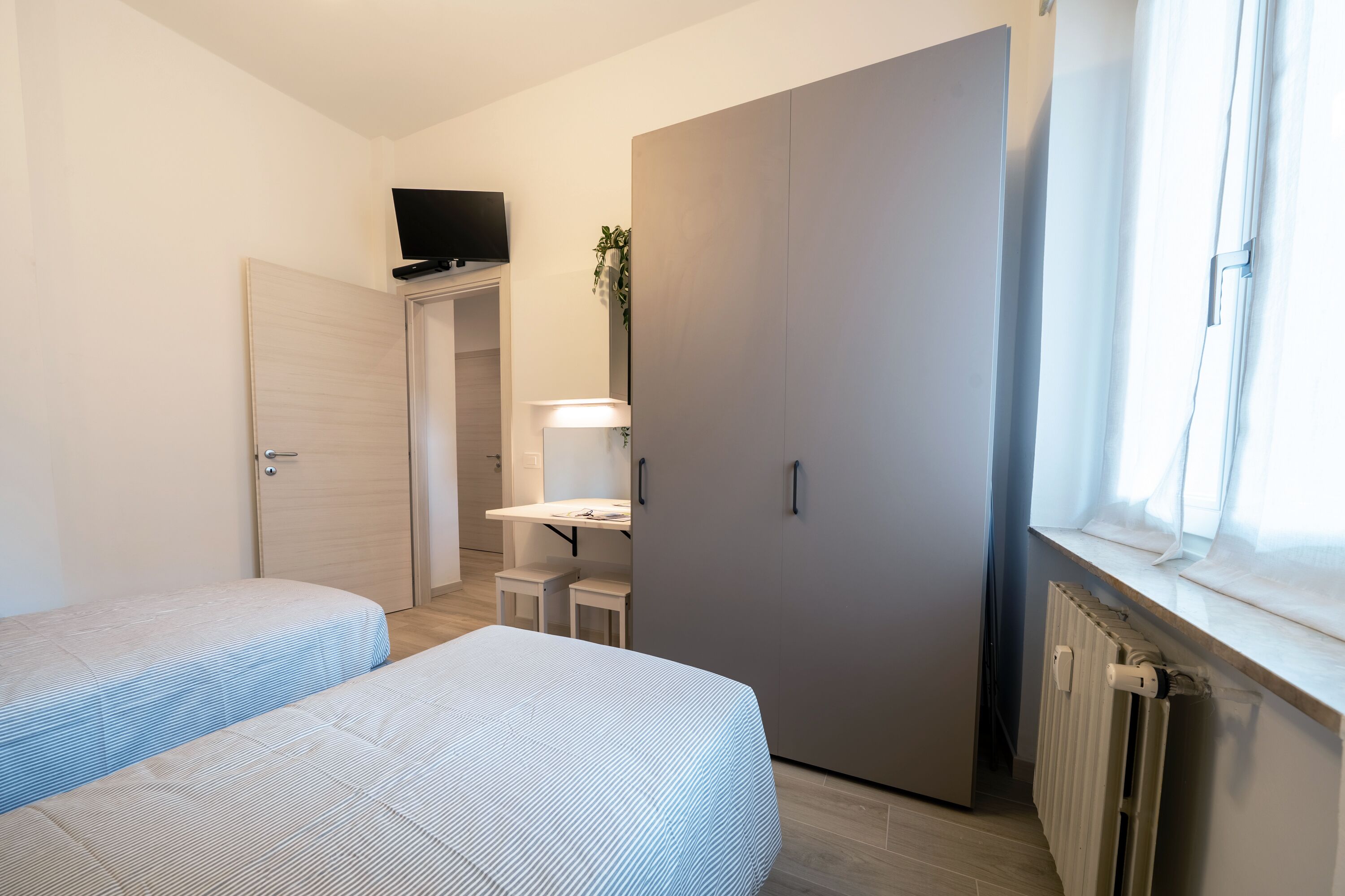 Une chambre avec deux lits, un bureau, une armoire, une télévision et une fenêtre avec des rideaux.