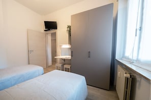 Ein Schlafzimmer mit zwei Betten, einem Schreibtisch, einem Kleiderschrank, einem Fernseher und einem Fenster mit Vorhängen.