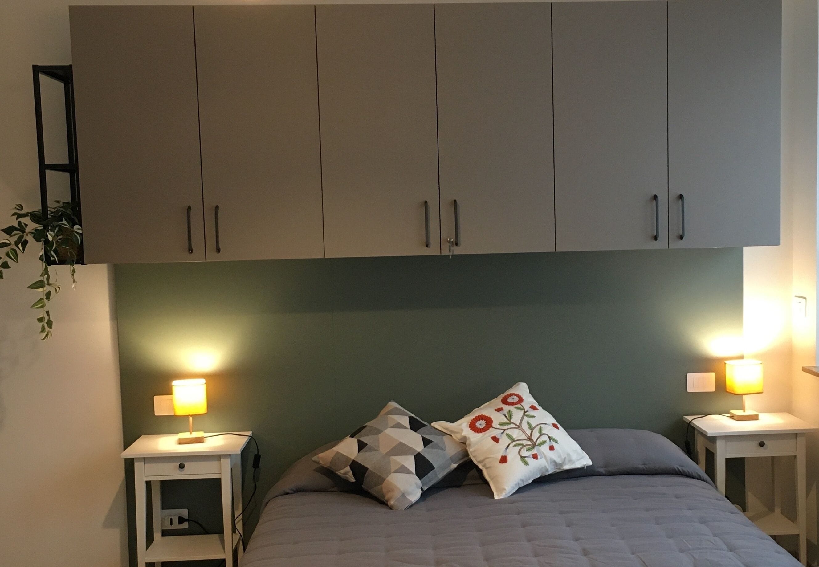 A bedroom with a bed, two bedside tables, a nightstand, a wall-mounted shelf, and a cabinet above the bed.