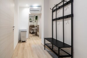 Ein Flur mit einer schwarzen Metallbank, einer weißen Mülltonne und einem im Hintergrund sichtbaren Badezimmer.